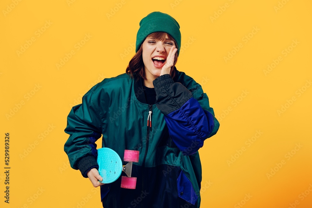an enthusiastic, emotional woman stands on a yellow background in a fashionable jacket and hat from the 90s and holding a skateboard shouts loudly into the camera an enthusiastic, emotional woman stands on a yellow background in a fashionable jacket and hat from the 90s and holding a skateboard shouts loudly into the camera