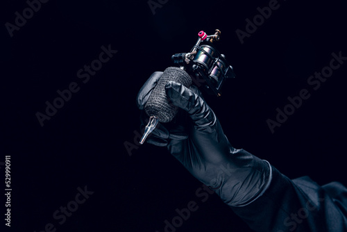 Machine for tattoo. Human hand in black protective glove holding professional equipment for making tattoo on body isolated on dark background. © master1305