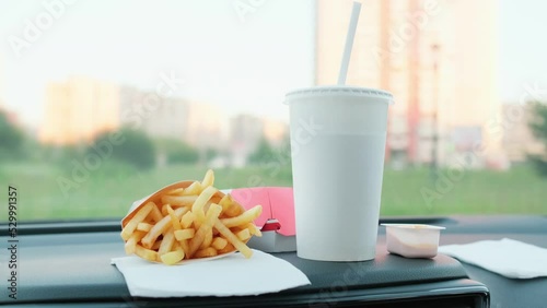 Fast food on a car dash. Burger, french fries and drink with straw in a car. Blurred cityscape on background.