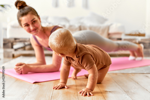   Mother and baby do sports together. The baby is crawling, and the mother is doing an exercise on the pink mat.