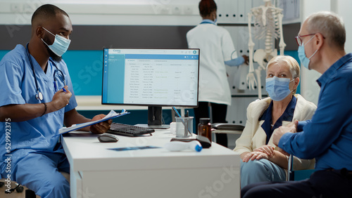 Medical worker consulting old woman with physical disability, doing checkup examination at clinic. Nurse talking to paralyzed patient and husband with face masks at appointment.