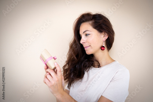 pretty young woman with beautiful long hair is holding a small christmas gift in her hand and has earrings that look like christmas tree baubles standing in front of light brown background