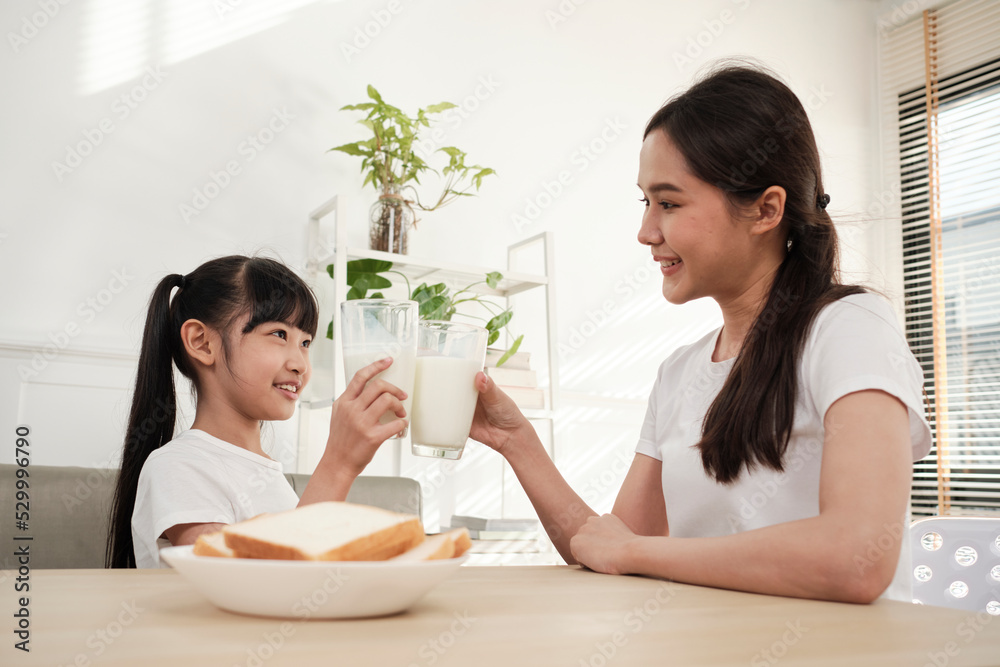 A Healthy Asian Thai family, little daughter, and young mother drink fresh white milk in glass and bread joy together at a dining table in morning, wellness nutrition home breakfast meal lifestyle. A Healthy Asian Thai family, little daughter, and young mother drink fresh white milk in glass and bread joy together at a dining table in morning, wellness nutrition home breakfast meal lifestyle.