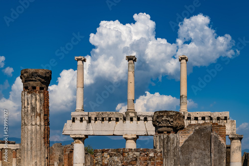 Historic remains of basilica and forum in destroyed Pompeii Italy. Row of colums and clouds resembling factory chimneys. Sunny day in popular tourist sight and world heritage monument of roman empire.