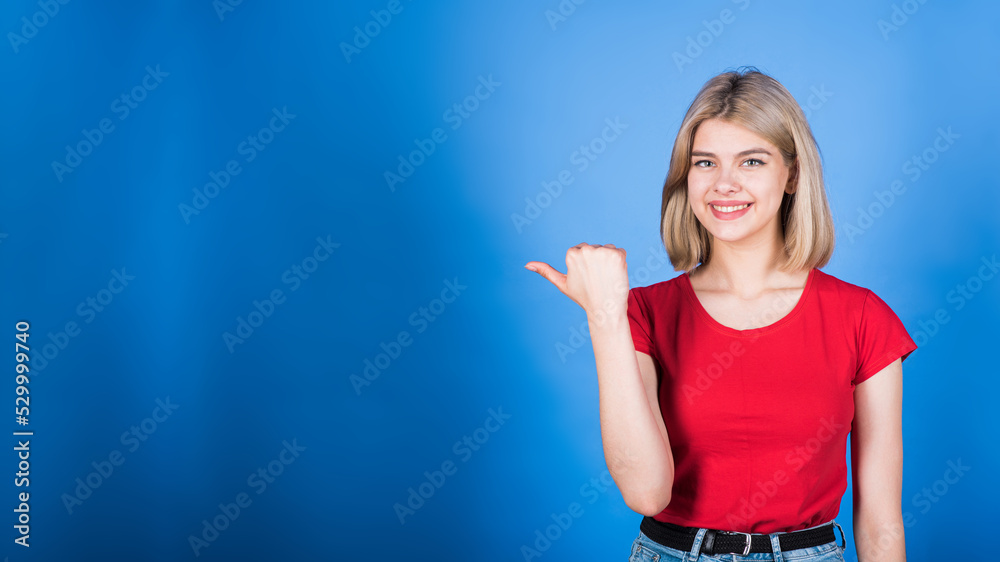 Young and attractive blonde caucasian girl in casual clothes pointing her thumb at copy space for advertising isolated on blue studio background.