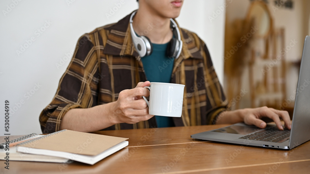 Asian male college student or freelancer working on his project on laptop while sipping coffee Asian male college student or freelancer working on his project on laptop while sipping coffee
