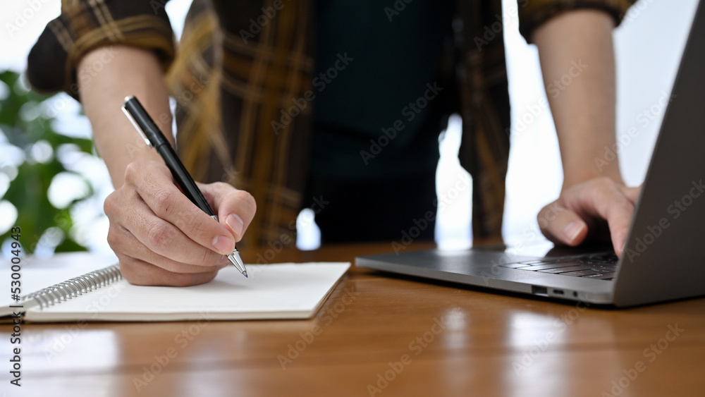 A professional male office worker in flannel shirt working at his desk, taking notes and using lapto A professional male office worker in flannel shirt working at his desk, taking notes and using lapto