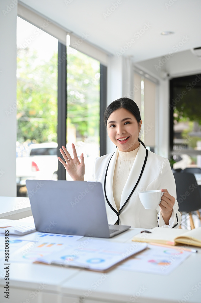 Portrait, Attractive Asian businesswoman sits at her office desk, sipping coffee and waving hand Portrait, Attractive Asian businesswoman sits at her office desk, sipping coffee and waving hand