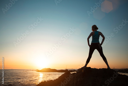 A woman does gymnastic yoga by the ocean in the setting sun.
