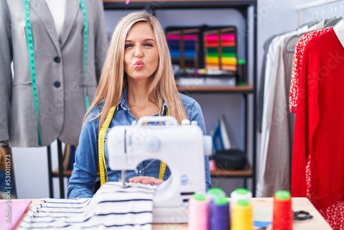 Blonde woman dressmaker designer using sew machine looking at the camera blowing a kiss on air being lovely and sexy. love expression.