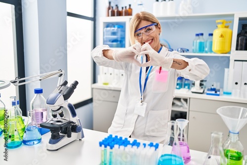Middle age blonde woman working at scientist laboratory smiling in love doing heart symbol shape with hands. romantic concept.