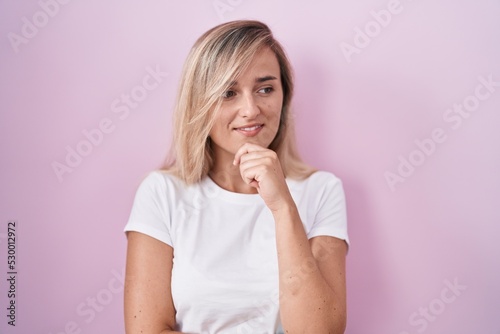 Young blonde woman standing over pink background with hand on chin thinking about question, pensive expression. smiling and thoughtful face. doubt concept.