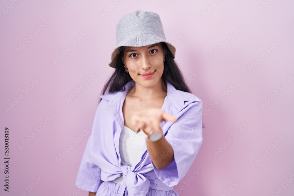 Young hispanic woman standing over pink background wearing hat smiling cheerful offering palm hand giving assistance and acceptance. Young hispanic woman standing over pink background wearing hat smiling cheerful offering palm hand giving assistance and acceptance.