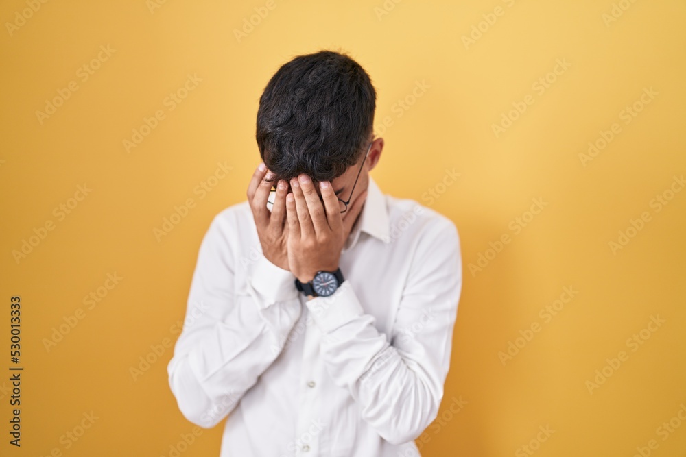 Young hispanic man standing over yellow background with sad expression covering face with hands while crying. depression concept. Young hispanic man standing over yellow background with sad expression covering face with hands while crying. depression concept.