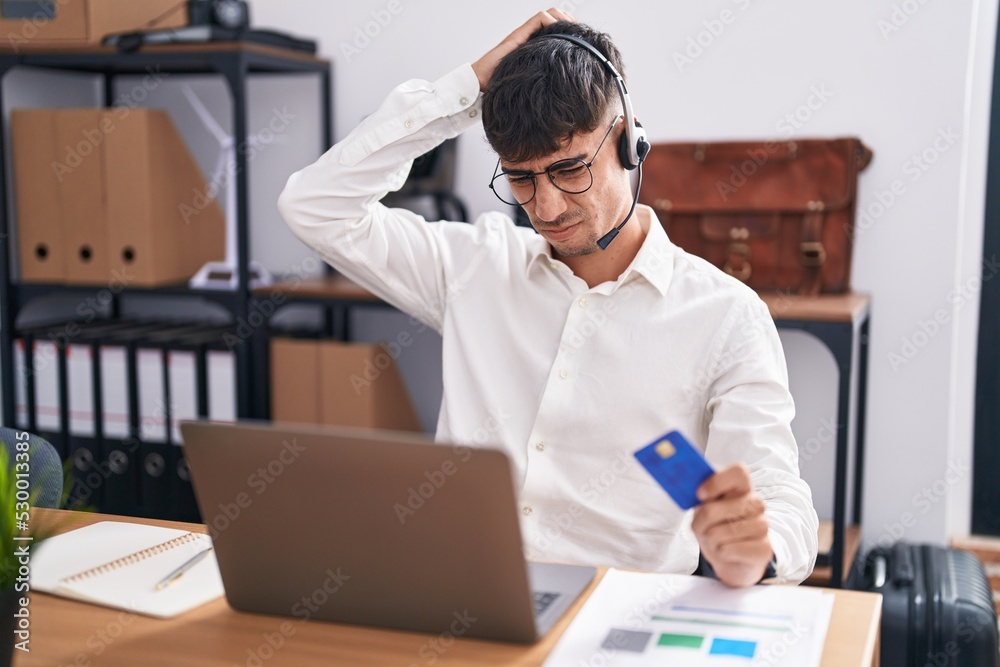 Young hispanic man working using computer laptop holding credit card confuse and wondering about question. uncertain with doubt, thinking with hand on head. pensive concept. Young hispanic man working using computer laptop holding credit card confuse and wondering about question. uncertain with doubt, thinking with hand on head. pensive concept.