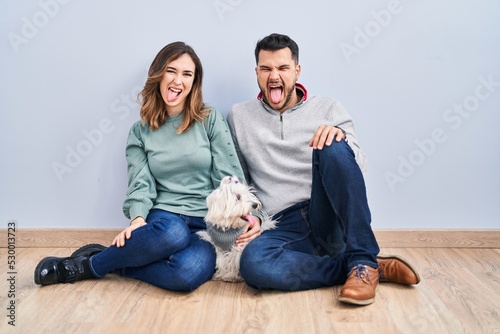 Young hispanic couple sitting on the floor with dog sticking tongue out happy with funny expression. emotion concept.