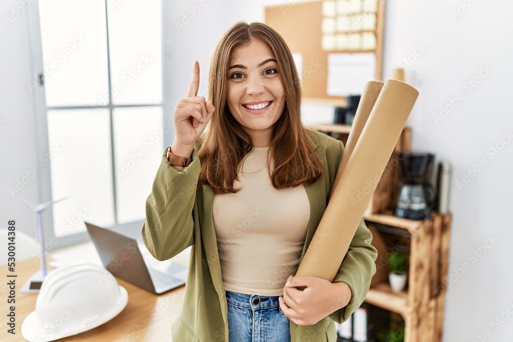 Young brunette woman holding paper blueprints at the office surprised with an idea or question pointing finger with happy face, number one Young brunette woman holding paper blueprints at the office surprised with an idea or question pointing finger with happy face, number one