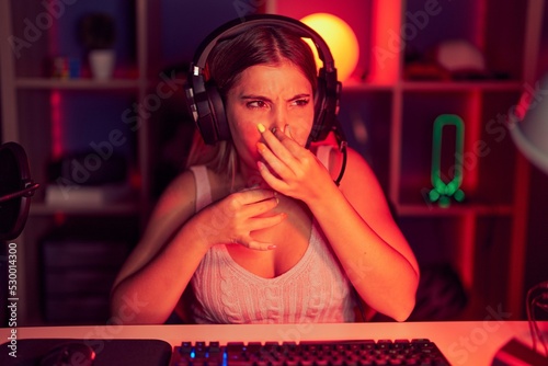 Young blonde woman playing video games wearing headphones smelling something stinky and disgusting, intolerable smell, holding breath with fingers on nose. bad smell