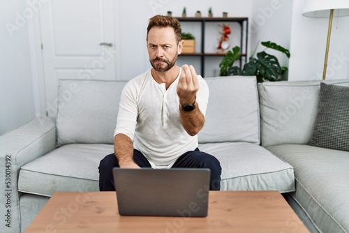 Middle age man using laptop at home doing italian gesture with hand and fingers confident expression