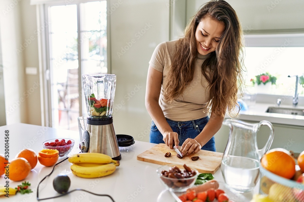 Young beautiful hispanic woman preparing vegetable smoothie with blender cutting date at the kitchen Young beautiful hispanic woman preparing vegetable smoothie with blender cutting date at the kitchen