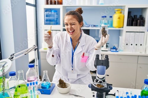 Young hispanic woman doing weed oil extraction at laboratory winking looking at the camera with sexy expression  cheerful and happy face.