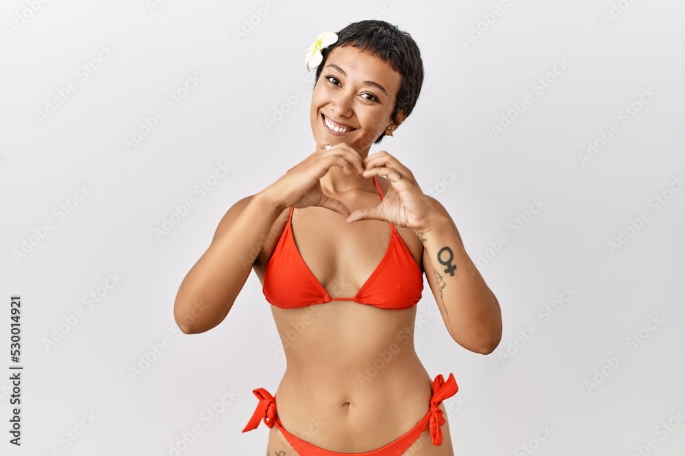 Naklejka premium Young hispanic woman with short hair wearing bikini smiling in love doing heart symbol shape with hands. romantic concept.