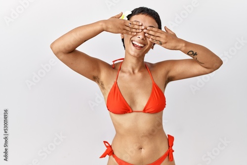 Young hispanic woman with short hair wearing bikini covering eyes with hands smiling cheerful and funny. blind concept.