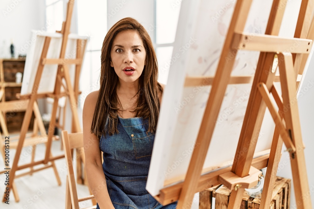 Young latin artist woman painting at art studio scared and amazed with open mouth for surprise, disbelief face