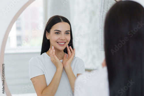 Portrait of a young beautiful brunette woman in a white T-shirt looking in the mirror at home, touching her face, skin, smiling, admiring.