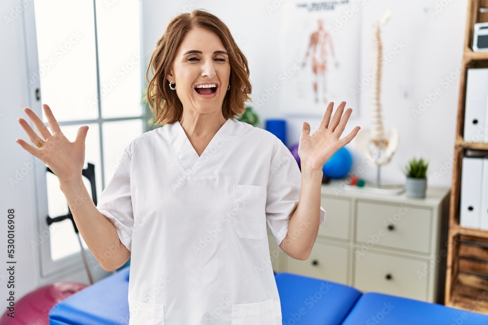 Middle age physiotherapist woman working at pain recovery clinic celebrating crazy and amazed for success with arms raised and open eyes screaming excited. winner concept Middle age physiotherapist woman working at pain recovery clinic celebrating crazy and amazed for success with arms raised and open eyes screaming excited. winner concept