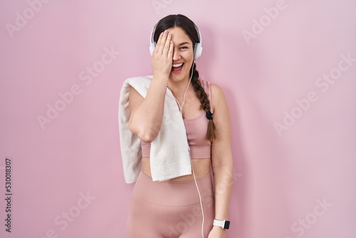 Young brunette woman wearing sportswear and headphones covering one eye with hand, confident smile on face and surprise emotion.