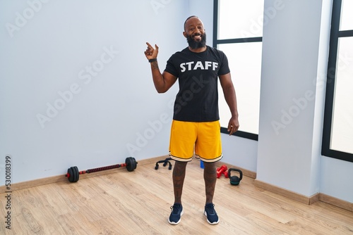African american man working at fitness gym smiling cheerful pointing with hand and finger up to the side