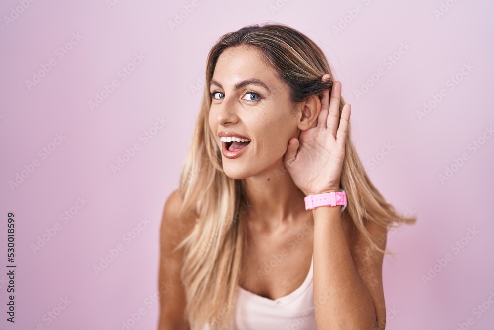 Young blonde woman standing over pink background smiling with hand over ear listening an hearing to rumor or gossip. deafness concept. Young blonde woman standing over pink background smiling with hand over ear listening an hearing to rumor or gossip. deafness concept.