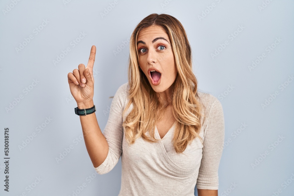 Young blonde woman standing over isolated background pointing finger up with successful idea. exited and happy. number one. Young blonde woman standing over isolated background pointing finger up with successful idea. exited and happy. number one.