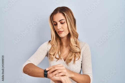 Young blonde woman standing over isolated background checking the time on wrist watch, relaxed and confident