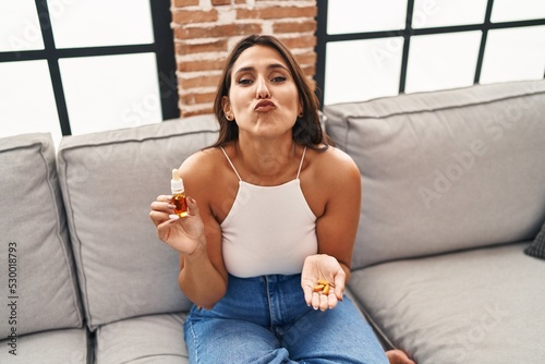 Young hispanic woman holding pills and cbd oil looking at the camera blowing a kiss being lovely and sexy. love expression.