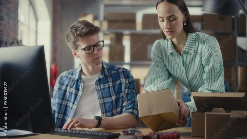 Two Employees Preparing Orders Made from Online Sales in Their Internet Shop. Man and Female Working in a Storeroom. Young Man Using Desktop Computer, Woman Confirming Contact Details on Boxes. Two Employees Preparing Orders Made from Online Sales in Their Internet Shop. Man and Female Working in a Storeroom. Young Man Using Desktop Computer, Woman Confirming Contact Details on Boxes.