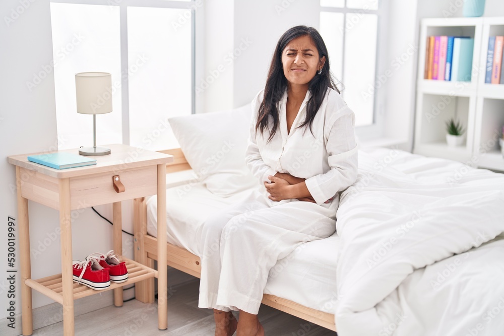 Young beautiful hispanic woman suffering for menstrual pain sitting on bed at bedroom Young beautiful hispanic woman suffering for menstrual pain sitting on bed at bedroom