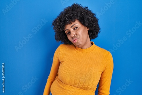 Black woman with curly hair standing over blue background looking at the camera blowing a kiss on air being lovely and sexy. love expression.