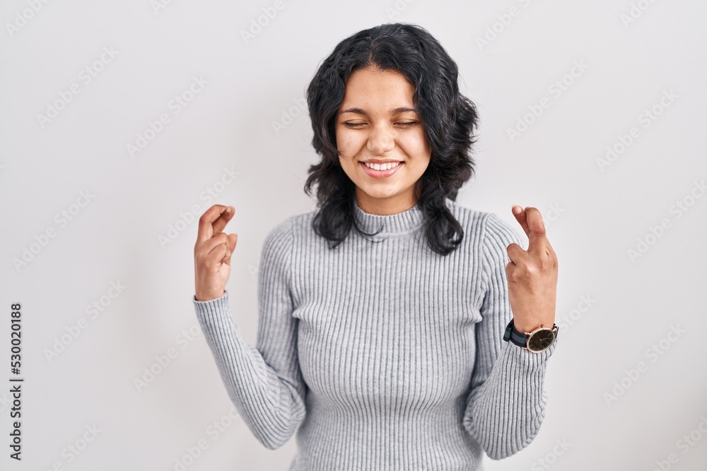 Hispanic woman with dark hair standing over isolated background gesturing finger crossed smiling with hope and eyes closed. luck and superstitious concept.