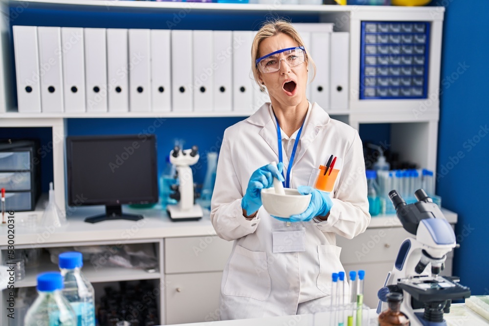 Young caucasian woman working at scientist laboratory in shock face, looking skeptical and sarcastic, surprised with open mouth