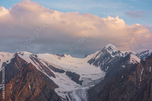 Dramatic aerial view to high snow mountain peak in early morning at dawn. Awesome scenery with sunlit snow mountains in cloudy sky at sunrise. Scenic landscape with large glacier in sunrise colors. © Daniil