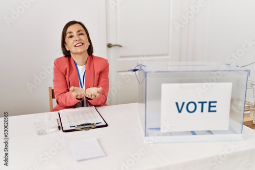 Beautiful middle age hispanic woman at political election sitting by ballot smiling with hands palms together receiving or giving gesture. hold and protection