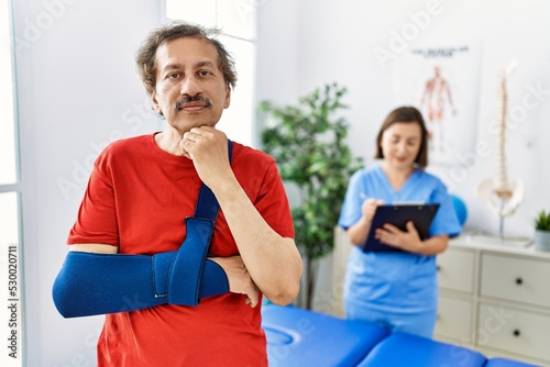 Southeast asian man wearing wearing arm on sling at rehabilitation clinic serious face thinking about question with hand on chin  thoughtful about confusing idea