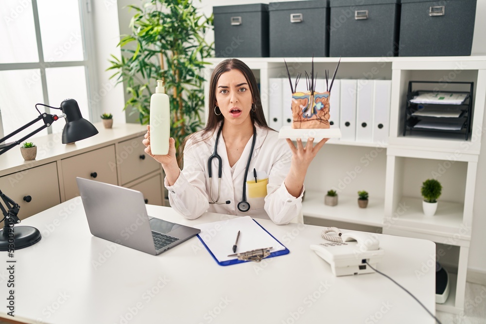 Hispanic doctor woman holding model of human anatomical skin and hair in shock face, looking skeptical and sarcastic, surprised with open mouth Hispanic doctor woman holding model of human anatomical skin and hair in shock face, looking skeptical and sarcastic, surprised with open mouth