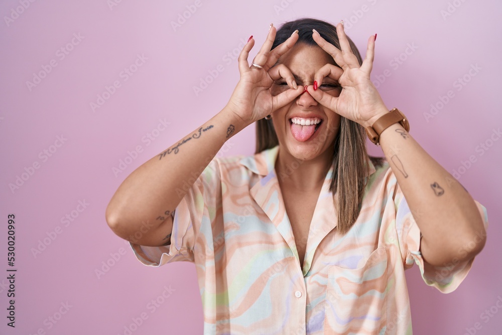 Blonde woman standing over pink background doing ok gesture like binoculars sticking tongue out, eyes looking through fingers. crazy expression. Blonde woman standing over pink background doing ok gesture like binoculars sticking tongue out, eyes looking through fingers. crazy expression.