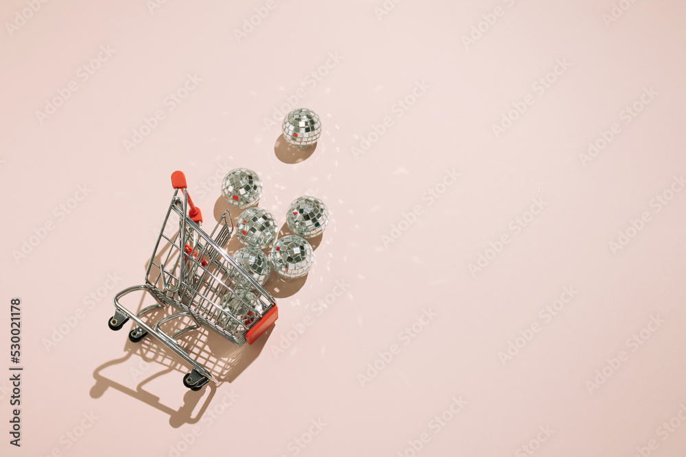 Overturned shopping cart with disco balls falling out on pink background.