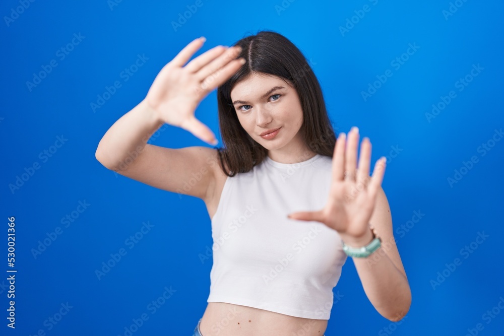 Fototapeta premium Young caucasian woman standing over blue background doing frame using hands palms and fingers, camera perspective