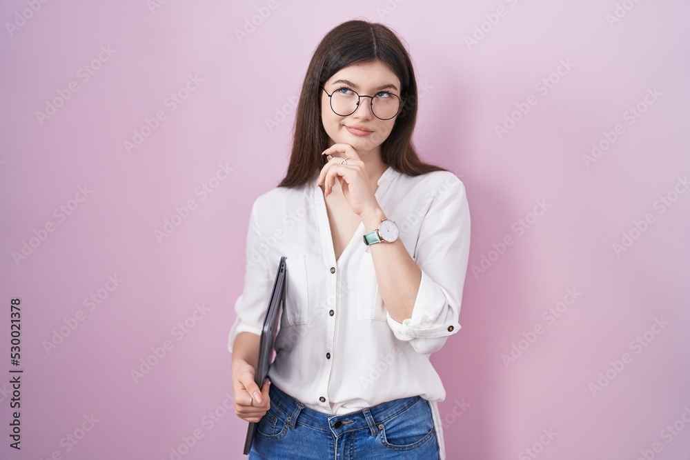 Young caucasian woman holding laptop with hand on chin thinking about question, pensive expression. smiling with thoughtful face. doubt concept.