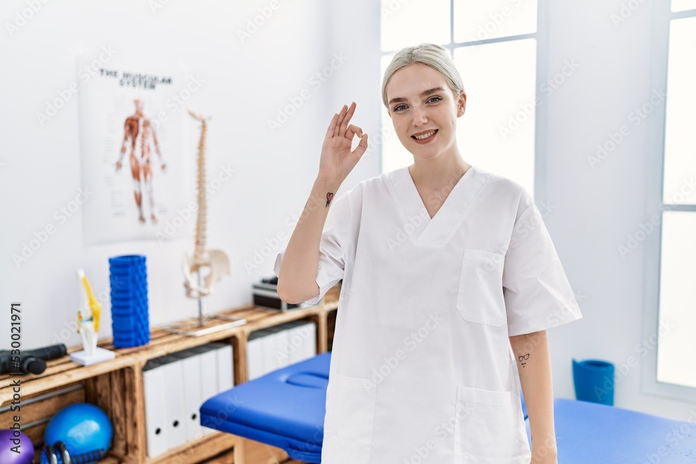 Fototapeta premium Young caucasian woman working at pain recovery clinic smiling positive doing ok sign with hand and fingers. successful expression.